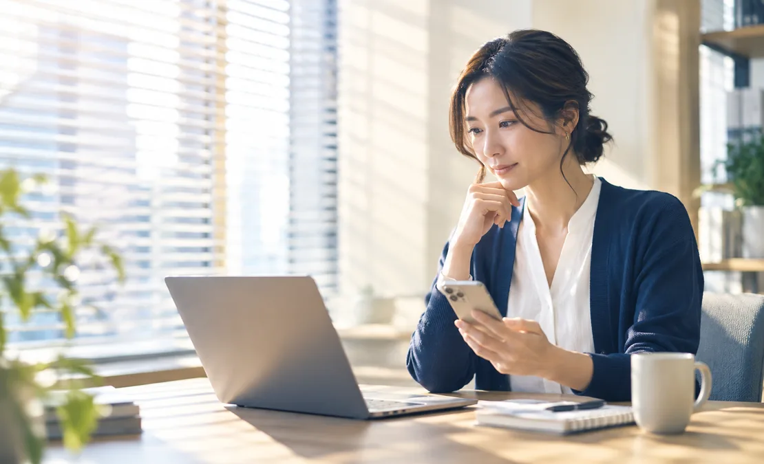 ECマーケターが窓際のデスクでノートPCとスマートフォンを見比べ、次の配信を考えている様子